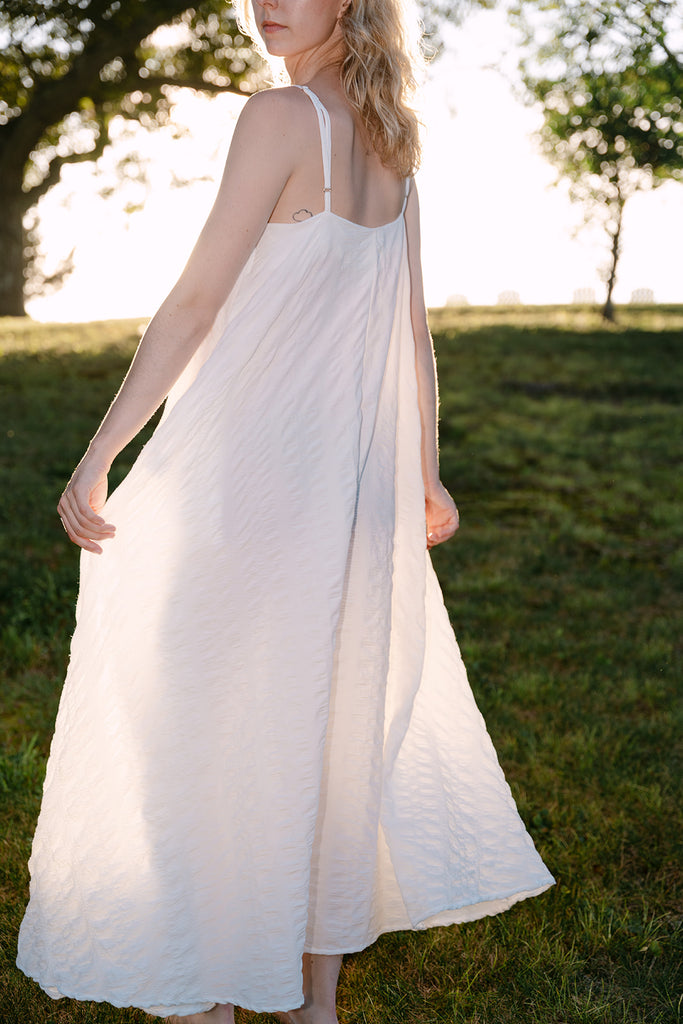 Woman wearing a white dress standing in a grassy field with trees in the background.